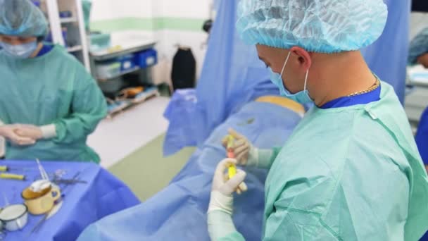 男医生拿着一对相互连接的注射器 专家把干细胞从一个注射器转移到另一个注射器 准备动手术的病人躺在手术台上 — 图库视频影像