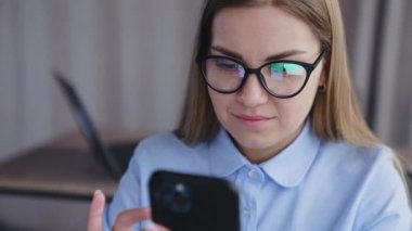 Sarı saçlı, gözlüklü, elinde telefon tutan ve araştıran bir kadın. Telefonuyla çalışan bir bayanın portresi. Kapatın. Yüksek açı görünümü.