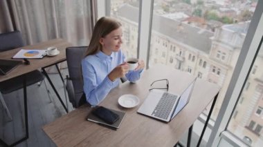 Kahve içen mutlu, dayanıklı genç bir kadın. Kadın dizüstü bilgisayarına bakıyor ve pencereden manzaranın tadını çıkarıyor. Yüksek açılı perspektif.