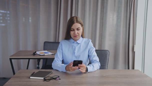 青いシャツを着ている長髪の女性が机に座っています 女性は彼女の携帯電話を見て 彼女の紙のノートにメモを取る — ストック動画