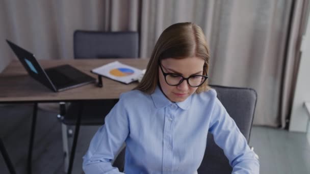 金发年轻的女雇员坐在桌旁 女人在她的笔记本电脑上工作 沉思地凝视着窗户 高角度视图 — 图库视频影像
