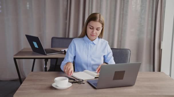 若い女性はオフィスで仕事で忙しい 女性はラップトップで作業し 彼女の紙のノートにメモを取る — ストック動画