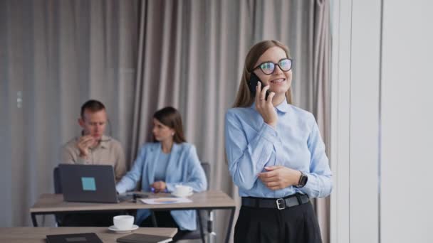 金发女人站在窗边的电话里说话 两名同事坐在笔记本电脑前的办公桌前 — 图库视频影像