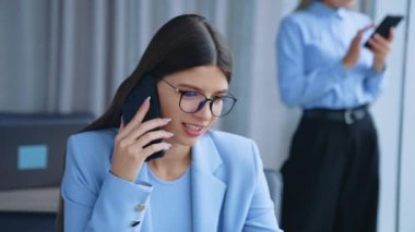 Gözlüklü, gülümseyen genç bayan masada oturuyor. Sarışın iş arkadaşı pencerede durup akıllı telefondan birine mesaj atıyor..