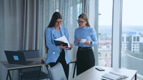 2人の女性同僚がオフィスの窓の近くに立っている オフィスの従業員は文書を見る仕事の問題を議論します 室内オフィスバックドロップ — ストック動画
