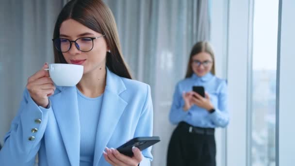 美しい黒髪の若い女性は 携帯電話を見ながらコーヒーを飲むメガネをかけている 女性同僚がバックドロップでスマートフォンで誰かにメッセージを送る — ストック動画