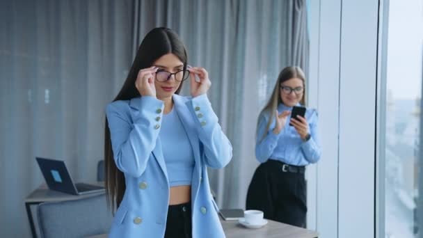 青いジャケットを着ている見栄えの良い若い髪の女性は オフィスの机に立っています ビジネス女性はカメラに笑顔でメガネをかけます バックドロップで窓に立って携帯を保持している別の女性 — ストック動画