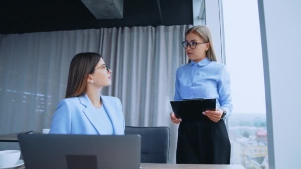 オフィスで話している女性社員 ドキュメントを保持するウィンドウに立っているブロンドの女性は机に座っている彼女の同僚に話す — ストック動画