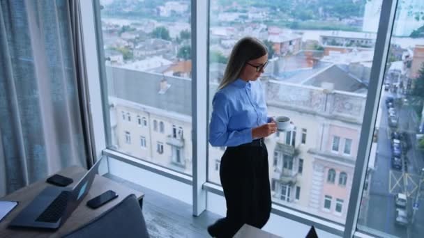 窓から美しい眺めのモダンなオフィスでのランチブレイク ブロンドの女性従業員は コーヒーのカップで窓まで歩いています 高角度ビュー — ストック動画