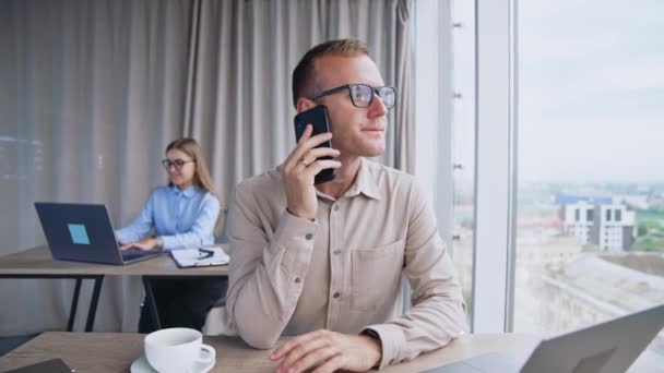 彼の耳元に電話をかけている男は 答えを待っている 男性従業員はスマートフォンを不満にしている 女性同僚がバックドロップで彼女のラップトップに印刷 — ストック動画