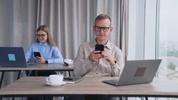 オフィスで電話をする 男はスマートフォンで笑顔で話す クローズアップ バックドロップの女性は一度に電話とラップトップを使用します — ストック動画