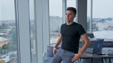 Gündelik giysiler giyen milenyum erkeği masaya yaslanıyor. Genç adam ellerini göğsünün üstüne koyup kameraya bakıyor. Portreyi kapat.