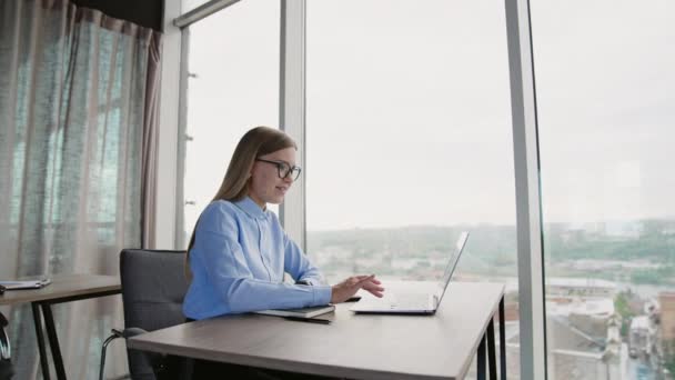 デスクに座っているコンピュータで働くメガネのブロンドの長髪の女性 パノラマウィンドウ付きのライトオフィスの背景 — ストック動画