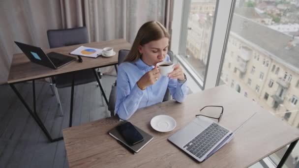 オフィスでランチ休憩でコーヒーを飲む 長髪の女性は ノートパソコンで机に座って窓を見ているコーヒーを楽しんでいます 高角度ビュー — ストック動画