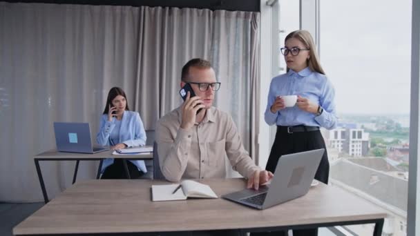 在办公室工作的人员工在工作时使用电话和手提电脑 女同事站在窗边喝咖啡 — 图库视频影像