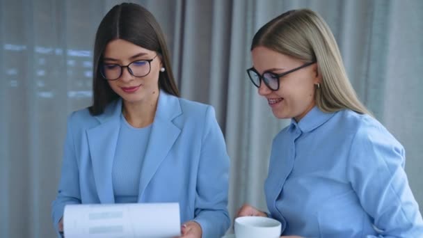 2人の女性の同僚が 彼らについてコメントする文書を調べた ブロンドの女性は彼女の手にカップを持っています オフィスバックドロップ — ストック動画