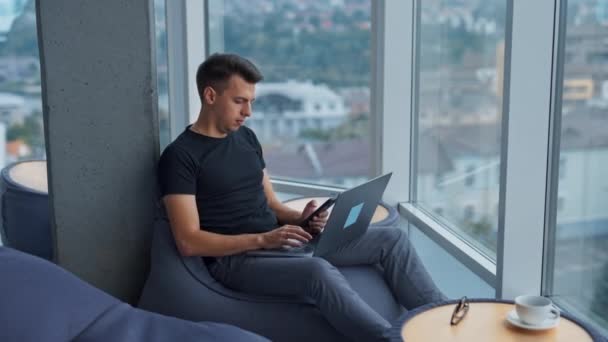年轻人坐在豆袋椅子上 膝上抱着笔记本电脑 商人手里拿着电话 城市景观在模糊的背景下 — 图库视频影像