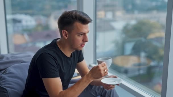 一个英俊的年轻人坐在大窗户旁的豆袋椅子上 男人一边喝着咖啡一边若有所思地看着窗户 模糊的背景 — 图库视频影像