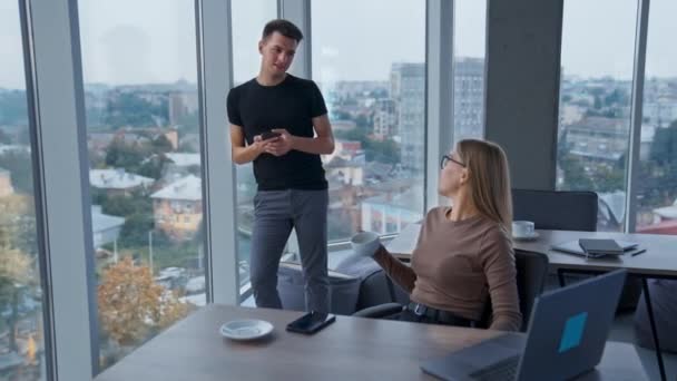 若い同僚がオフィスで話している デスクに座ってコーヒーを飲んでいる女性 バックドロップのパノラマウィンドウ付きオフィス — ストック動画