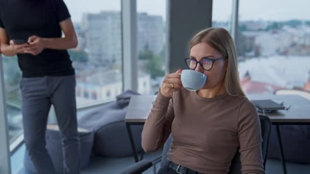 ブロンドリラックスした女性は 同僚と話すコーヒーを飲むメガネを着ています 窓の後ろに立っている若い男が携帯電話を持っている バックドロップのシティスケープ — ストック動画