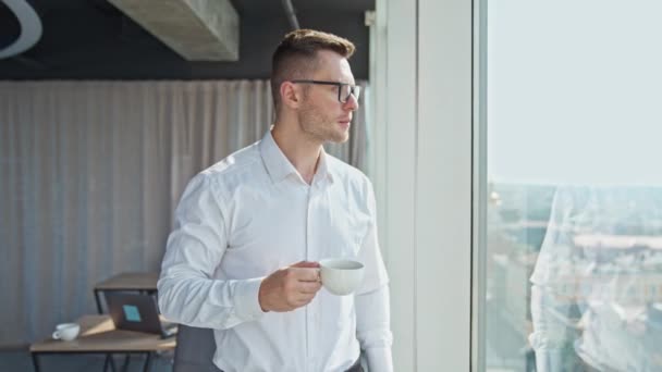 ライトオフィスに立っている若い自信あるビジネスマン 白いシャツの男がパノラマウィンドウを眺めながらコーヒーを飲む — ストック動画