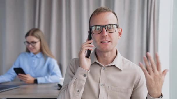 中年ハンサムな男性が手でジェスチャーする電話でメガネをかけています バックドロップの女性同僚は 携帯電話を見ているラップトップの前に机に座って — ストック動画