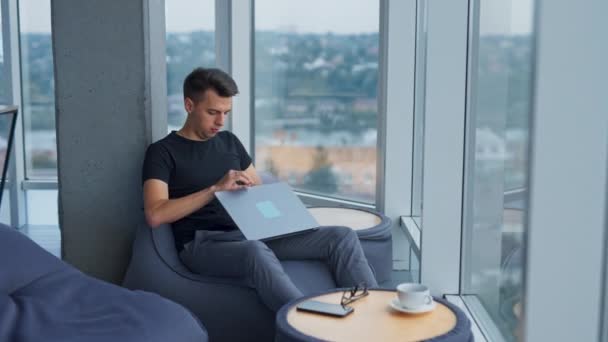 ビーン バッグの椅子に座っている若い実業家がノートパソコンを開けます 大きなパノラマウィンドウの近くの居心地の良いオフィスで仕事をしている忙しい男 — ストック動画