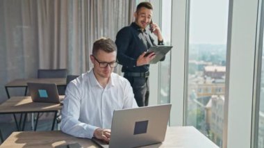 Geniş ışıklı odada orta yaşlı genç ofis çalışanları. Beyaz gömlekli bir erkek dizüstü bilgisayarda çalışıyor ve arkasındaki adam telefonla konuşuyor ve gülüyor..