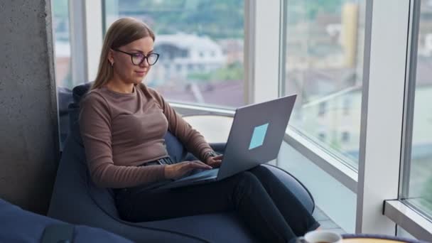 漂亮的金发女士坐在豆袋椅子上 在笔记本电脑上工作 女士一边工作一边看着她坐着的大玻璃窗 分散了注意力 — 图库视频影像