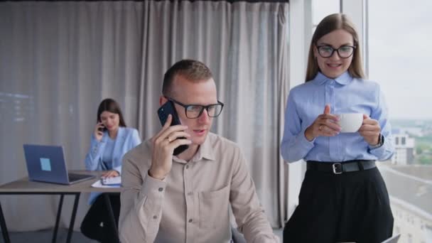 電話でのビジネス会話 電話でクライアントと話す男性と スクリーンで手札のカップの横に立っている女性 バックドロップの女性の同僚 — ストック動画