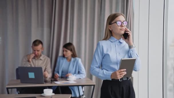 眼鏡をかけたビジネスレディは 窓際のオフィスに立っている電話で話す 女性は手元に紙のノートを握っている バックドロップで働く同僚 — ストック動画