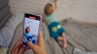 Anne, oğlunun akıllı telefondaki videosunu çekiyor. Küçük bebek yatakta emekliyor beyaz tuğla duvarı keşfediyor. Bulanık arkaplan.