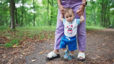 Küçük oğlana yürümeyi öğretiyorum. Anne bebeğini ellerinden tutuyor ve çocuğuna parkta yol boyunca önderlik ediyor. Doğa bulanık arkaplan.