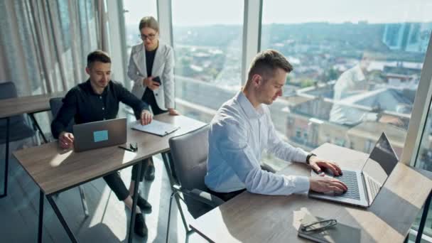 晴れたモダンなオフィスで働くオフィスの従業員 ノートパソコンの前に座っている男性 黒シャツの男性同僚の近くに立っている白いジャケットの女性 — ストック動画