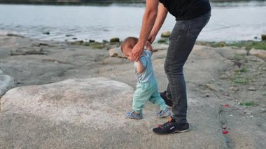 Kot giyen bir adam küçük oğlunu yürümeye zorluyor. Mavi elbiseli güzel erkek bebek taşlara basıyor. Nehir kıyısında..