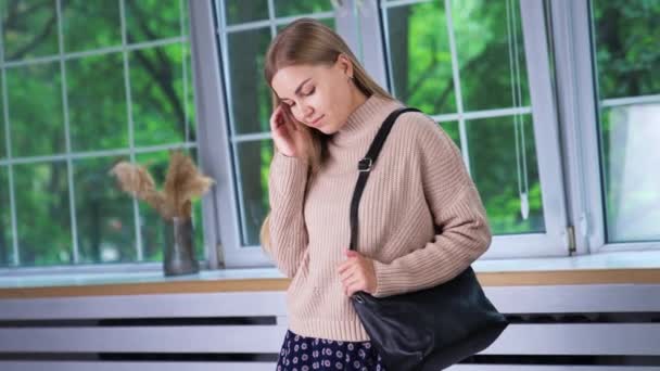 窓の屋内で立っているベージュセーターの若い金髪の女性 彼女の肩に黒いバッグを持っている美しい女性 — ストック動画