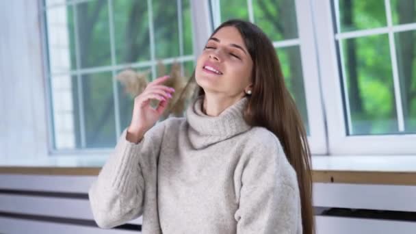 屋内暖かいセーターで魅力的なブルネット 髪に触れた若い女性は カメラに向かってポーズをし 笑顔を浮かべた 現代の服と履物プレゼンテーション — ストック動画