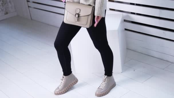 暖かいセーターとグレーバッグを保持するブラックパンツのモデル 屋内でファッショナブルな靴を披露する女性 ホワイトバックドロップ — ストック動画
