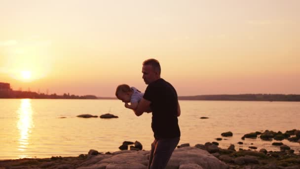 父親を愛し 子供と時間を過ごした 父親は息子を腕に抱きしめ 投げつけている バックドロップの川の上の夕日 — ストック動画
