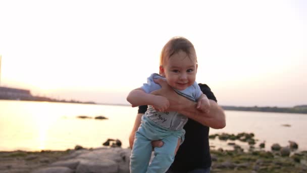 父親は海岸で息子と遊んでいました かわいい赤ん坊の少年は 父親の手でカメラに近づいている バックドロップの空をクリア — ストック動画