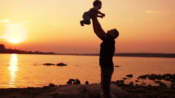 小心爸爸和他的孩子在河边玩慈爱的父亲抛掷他的儿子和孩子高兴 夕阳西下 — 图库视频影像