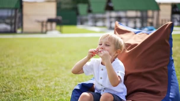 5歳の少年が屋外に座っている顔をしています 明るい太陽の下でビーン バッグの椅子で休むことを楽しんでいる小さな子供 クローズアップ — ストック動画