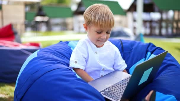 ハッピーな小さな白人少年がコンピュータで何かを見ている ブロンドの子供は 自然の中で椅子に座って幸せに微笑んでいます ブラーレッド バックドロップ — ストック動画