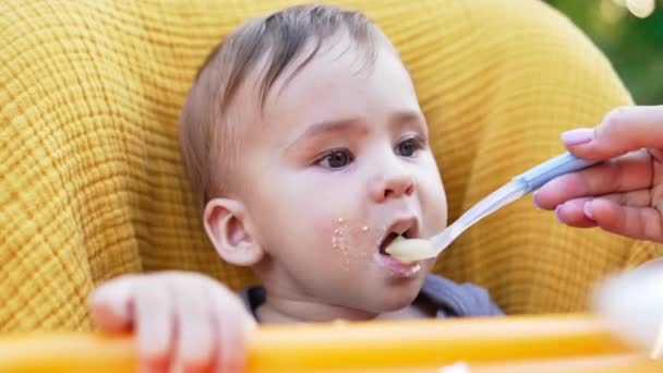 スプーンからの赤ちゃんの栄養 かわいいコーカサス人の子供は お粥を食べながら顔を汚した クローズアップ ブラーレッド バックドロップ — ストック動画