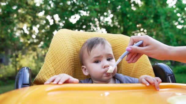 屋外の黄色い椅子に座っている愛らしい赤ん坊の少年 母は子供にフルスプーンを与え 彼は食べる 輝かしい自然の背景 — ストック動画