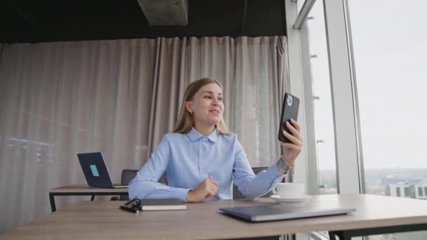 一位穿着蓝色衬衫的漂亮女士在前面的电话里看着她 微笑的女人通过在线视频聊天聊天 窗口背景下的城市景观 — 图库视频影像