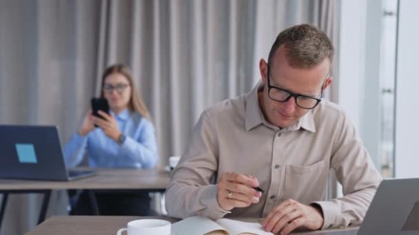 メガネをかけている忙しい男性の従業員は 紙のノートの上に頭を曲げました バックドロップで彼女の携帯を笑顔で見た女性 — ストック動画