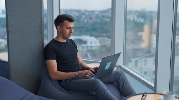 年轻的男性自由职业者在笔记本电脑上工作 舒适地坐在全景窗前 侧视图 城市景观在模糊的背景下 — 图库视频影像