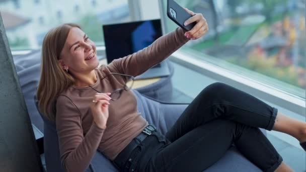 一个面带微笑的年轻女人半躺在豆袋椅子上 手里拿着电话 女士在休息的时候有视频聊天 模糊的背景 — 图库视频影像