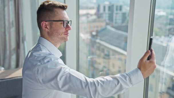 一位中年男子 穿着白衬衫 戴着眼镜 在一间阳光灿烂的办公室里 商人在智能手机上进行视频聊天 侧视图 — 图库视频影像